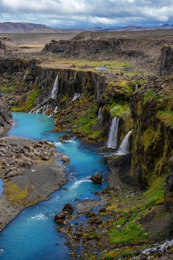 Turkuaz nehri, İzlanda, Studlagil 'de küçük şelaleleri olan bazalt kanyon boyunca esiyor. Yosun tutmuş kaya duvarları ve seyrek dağlık arazi bitkileri bulutlu gün ışığında görünür..