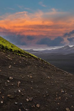 Çerçeveyi kaplayan koyu kül ve çakıl taşları, yosunlar sırtı kaplıyor, buzullar puslu bir şekilde kayboluyor, ve akşam ışığı İzlanda 'da koyu mavi bulutları turuncu ve pembeye boyuyor..