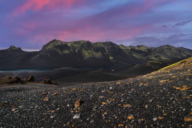 Tırtıklı yosunlu dağlar, İzlanda 'nın Landmannalaugar yakınlarındaki dağlık arazilerinde siyah kum ve çakıl taşları, sarı çakıl taşları ve alacakaranlıkta soluk bir buzul gölüyle yükselir..
