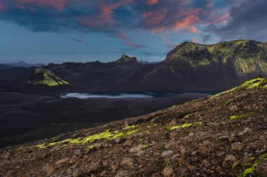 Karanlık lav tarlaları ve kül ovaları mavi bir buzul gölü oluşturur. Yosun, İzlanda 'nın yüksek dağlarında alacakaranlıkta süzülür..