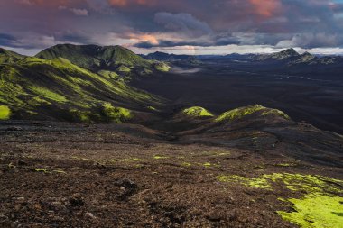 İzlanda 'nın Landmannalaugar yakınlarındaki dağlık bölgelerinde kara ovalar, açık yeşil yosunlar, lav tarlaları ve fırtına bulutları dağılırken kar zirveleri, geniş bir vadi boyunca uzanan ılık ışık bulutları görülüyor..