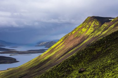 Landmannalaugar yakınlarındaki İzlanda 'da volkanik dağları kaplayan eğimli bir yosun, alçak bulutlar ve fırtınalı ışıkların altında çelik mavi göllere kadar uzanan tepeleri olan güneş ışığını yakalar..