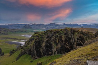 İzlanda 'da gün batımında siyah kum izleri ve karlı zirvelerle örülmüş nehirler yeşil bir vadide zikzak çizerken ziyaretçiler bazalt bir uçurumun tepesinde duruyorlar..