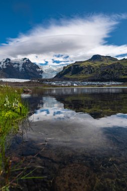 İzlanda 'da merceksi bir bulut, yosunlu dağlar ve ön planda çimenler ve taşlar bulunan mavi beyaz bir buzulun gündüz görüşü.