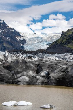 Kül bantlı bir İzlanda buzulu, sütlü bir gölde sürüklenen buzdan buzul, yeşil siyah volkanik dağlar ve parlak bir buz örtüsü..