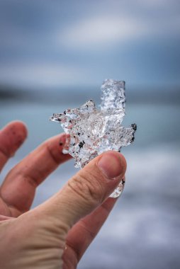 Bir el, açık mavi denize karşı uzanan ve Elmas Sahili yakınlarındaki İzlanda sahilinde bulutlu bir ışığa karşı siyah kum parçacıkları olan yarı saydam bir buz parçasını tutuyor..