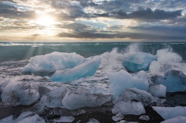 Pürüzlü mavi buz, Diamond Beach, Jokulsarlon, İzlanda 'da dalgalar çarptığında siyah kuma dayanır. Alçak güneş, öğleden sonra geç saatlerde Kuzey Atlantik 'e gümüş ışık saçar.
