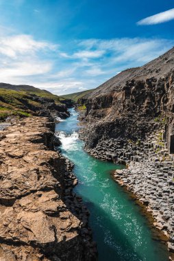 İzlanda, Studlagil 'deki altıgen bazalt sütunların arasından geçen turkuaz bir nehir. Gün ışığı, yüksek kontrast, çıtır çıtır ayrıntı, akıntı manzarası, çorak volkanik yamaçlar, uzak çağlayanlar..