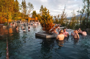 Ziyaretçiler gün batımında İzlanda 'daki bir jeotermal orman gölünde sıcak buhar altında dinleniyorlar. Katmanlı havuzlar, kaya kenarları, ahşap güverteler ve küçük adalar huş, çam, dağlar ve bir göl kenarında otururlar..