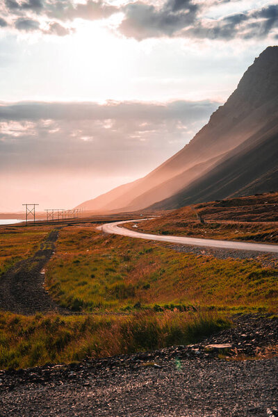 A curving coastal road threads past golden grass and a steep mountainside in Iceland, power lines trace the shore, mist softens layered peaks in late afternoon light.