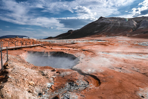 Steam rises from a bubbling mud pool as visitors follow a boardwalk at Hverir near Namafjall in North Iceland, midday light revealing ochre earth and fumaroles.