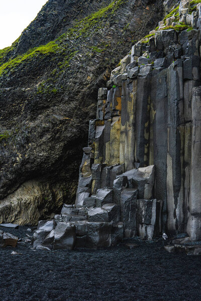 Базальтовые колонны поднимаются угловыми ступенями к черному песку на пляже Рейнисфьяра, Вик и Мирдал, Исландия. Мосс оттеняет рок, когда пасмурный свет раскрывает холодные серые шестиугольные формы.