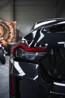 Yakından bakınca bir atölyede parlak siyah bir BMW M2 2025 görünüyor. LED arka lamba, çamurluk, ve tampon çerçeveyi doldurmak, arkasında kırmızı bir Ducati bulanık.