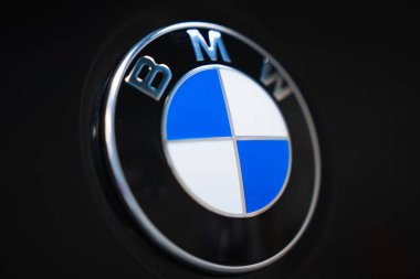 Yakın planda BMW yuvarlak amblemi bir atölyedeki araç panelinde görülüyor. Yumuşak ışık ve sığ alan derinliği vurgulanmış metal ve doygun renkler.