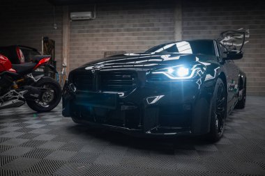 Parlak siyah bir BMW M2 2025 havalı bir atölyenin altında duruyor sağ farları açık, sola dönük, kırmızı bir Ducati spor motosikleti sol arka duvardaki garajda park ediyor..