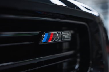 BMW M2 rozetini kapat ve atölyedeki parlak siyah ızgarada üç renkli şeritler kullan. Yumuşak ışık ve sığ alan derinliği metali ve modern havayı vurguluyor.