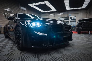 Parlak siyah bir BMW M2 2025 modern bir atölye bölümünde duruyor. Mavi LED farlar kareli zemine yansır. Ayrıntılı araçlar ve başka bir araç dengesiz bir hazırlık sahnesi ayarladı.