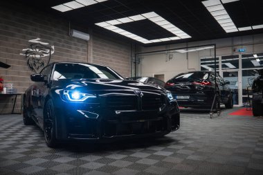 Pırıl pırıl, siyah bir BMW M2 mavi LED farlı modern bir atölyeye park edilmiştir. Zemin kareli, şebeke aydınlatma, tuğla duvarlar, aletler ve bir Ducati amblemi vardır..