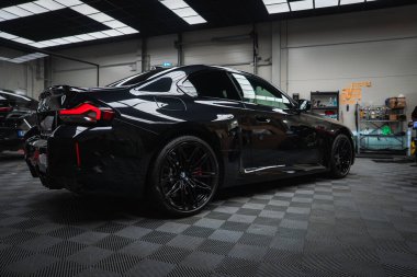 Parlak siyah bir BMW M2 2025, iyi aydınlatılmış bir atölyede aletler ve raflarla, LED L şeklindeki arka farlar parlıyor, dörtlü egzozlar ve parlak kemerler performans odaklarını gösteriyor