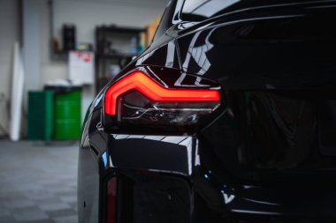 Bir atölyede, raflarda, aletlerde ve yeşil kutularda kırmızı parlayan BMW M2 2025 arka farını kapatın. Dağınık iç ışık parlak siyah bedene yansıyor.