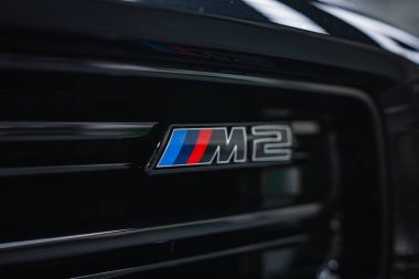 Yakından bakınca BMW M2 rozeti görünüyor. Kapalı alanda parlak siyah ızgara üzerinde M çizgileri, atölye ayarları, sığ alan derinliği, düşük aydınlatma yüzeyleri metal yüzeyler..