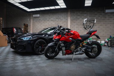 2025 BMW M2 ve kırmızı Ducati spor motosikleti gri tuğla duvarlı, kareli zemin döşemeli, LED panel ışıklandırmalı ve aslan arması amblemli bir atölyede oturuyorlar..