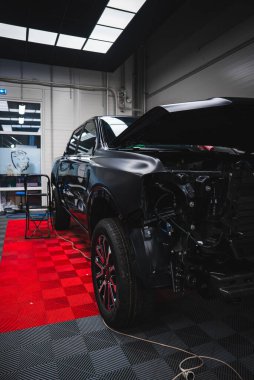 Parlak siyah BMW M2 2025 parlak paneller altında kırmızı ve siyah paspaslar üzerinde oturur, ön tampon ve ışıklar çıkarılır, kablolar görünür, servis arabası ve kablo aktif iş olduğunu gösterir.