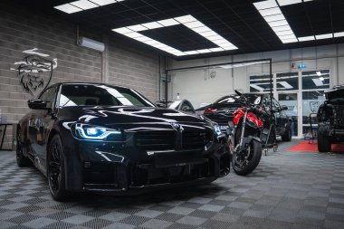 Parlak siyah bir BMW M2, kareli bir garaj zemininde, kırmızı ve siyah bir Ducati 'nin yanında, tuğla duvarları, cam bölme kapıları ve aslan logosu olan serin ızgara ışıklarının altında oturuyor..