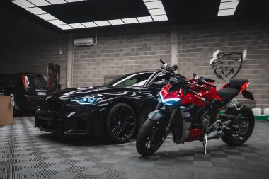 Parlak siyah BMW M2 2025 ve kırmızı Ducati endüstriyel bir atölyede yan yana durur, mavi LED farlar ve DRL 'ler yakılır, aslan amblemi ve aletler görünür, dama zemininde parlar.