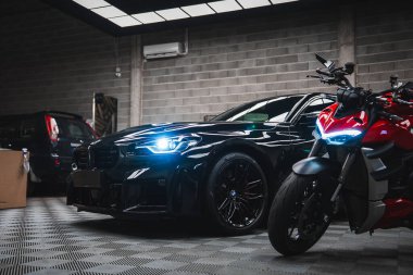 Pırıl pırıl siyah bir BMW M2 ve kırmızı bir Ducati kapalı alandaki düşük açılı yansıtıcı yüzeyler ve serin difüzyon ışıklarıyla aydınlatılan bir endüstriyel atölyede oturuyorlar..