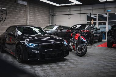Parlak siyah bir BMW M2 2025 ve kırmızı ve gümüş bir Ducati modern bir atölyede, şebeke ışıkları altında gri tuğla duvarlar, cam kapılar ve gece kalkan amblemi altında.