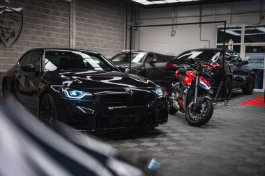 Parlak siyah bir BMW M2 ve kırmızı ve beyaz bir Ducati gri kareli bir garaj zemininde oturuyor, serin ışıkların altında geniş açılı, düşük manzaralı, mavi LED detaylar görünür.
