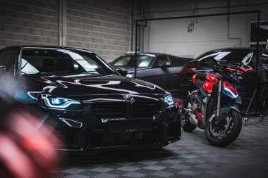 Çağdaş bir atölyede, beton duvarları, dama zemini ve koyu renkli arabaları olan kırmızı ve siyah bir Ducati 'nin yanında mavi farları olan parlak siyah bir BMW M2.