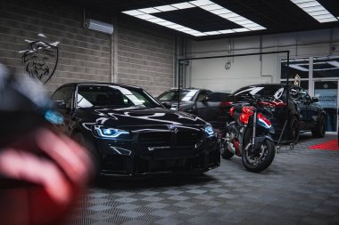 Parlak siyah bir BMW M2 ve kırmızı ve siyah bir Ducati gri tuğla duvarları, dama yeri, aslan arması logosu ve cam kapıları olan modern bir atölyede parlak panel ışıkları altında oturuyorlar..