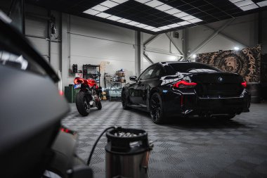 Parlak siyah BMW M2 2025 ve kırmızı Ducati kareli zemini, şebeke tavan lambaları, malzeme rafları ve görüş alanında bir vakum tüpü olan bir atölyede oturuyorlar..