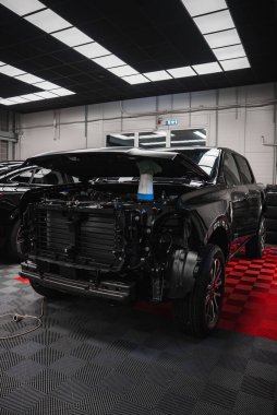 Siyah bir pikap parlak bir atölyede duruyor, kaput destekli, radyatör ve soğutucu açık, kareli zemin, istiflenmiş lastikler, şebeke ışıkları, onarım ve bakım çalışmaları devam ediyor.