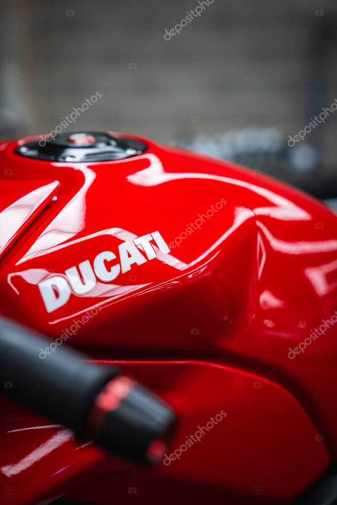 A tight close up shows the glossy red Ducati fuel tank and white logo, with sculpted contours and a hint of handlebar in muted indoor workshop lighting.