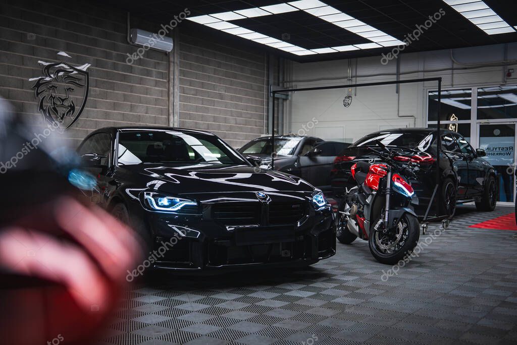 A glossy black BMW M2 and a red and black Ducati sit under bright panel lights in a modern workshop with gray brick walls, checker floor, lion crest logo, and glass doors.