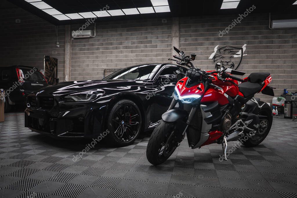 Glossy black BMW M2 2025 and red Ducati naked motorcycle sit indoors under bright panels, gray cinderblock walls, checker rubber floor, lion crest logo, AC units visible.