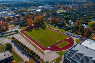 Hava görüntüsü Jelgava, Letonya 'da kırmızı bir pist ve futbol sahası gösteriyor. Dairelerin, bahçe arazilerinin ve güpegündüz sonbahar ağaçları ve şehir bloklarının olduğu bir spor kompleksinin yanında..