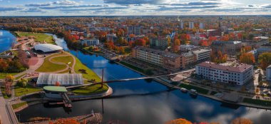 Litvanya 'nın Jelgava kentinin havadan görünüşü, Lielupe Nehri' nin yaya köprüsünün, parkların ve beyaz çatı spor kompleksinin yanından geçerken, gün ortası ışığı ve canlı sonbahar renginde