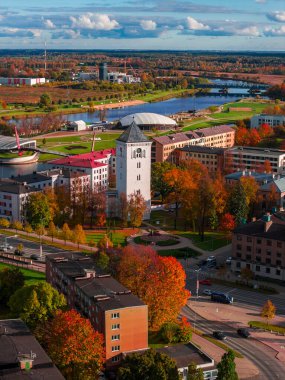Jelgava, Letonya 'nın öğleden sonra güneşinin havadan görünüşü. Beyaz bir saat kulesi ve kare kırmızı ve kehribar ağaçlarının arasında duruyor. Lielupe Nehri, köprüler ve kubbeli arena yakınlarda belirir..