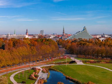 Riga, Letonya 'nın sonbaharın sonlarına doğru havadan bakıldığında altın ağaçlar, kıvrımlı bir park göleti ve yollar görülüyor. Ulusal Kütüphane ve Eski Kasaba Daugava Nehri boyunca yükseliyor..