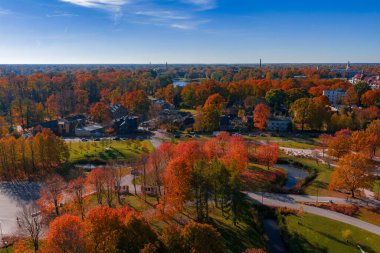 Riga, Letonya 'nın hava manzarası, evlerin etrafındaki sonbahar ağaçlarını, kıvrımlı bir park deresini ve gün ortasında ufukta bacaları ve kilise kuleleri olan küçük bir gölü gösteriyor..