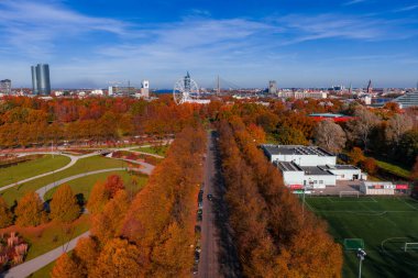 Riga, Letonya 'nın hava görüntülerinde, altın park caddeleri, beyaz dönme dolap, Vansu Köprüsü, nehir, kuleler, kilise kuleleri, spor kompleksi ve açık bir sonbahar günü sunumları görülüyor..