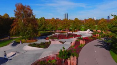 İnsanlar yürürken ve kaykay yaparken hava manzarası beton bir kaykay parkının ve Riga parkındaki patikaların üzerinde süzülüyor. İki silindirik yüksek bina ve bir dönme dolap bir sonbahar sahnesi..