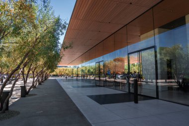 Yüzen ahşap çoraplarla kaplı cam duvarlı pavyon Cupertino, California 'daki Apple Park' ta bir gezinti güvertesinde. Gün ortası ışığı, az sayıda insanın girmesiyle aydınlık gölgeler oluşturur..