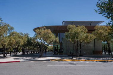 Cupertino 'daki Apple Park Ziyaretçi Merkezi cam bir pavyon ve ince kantilli çatı, zeytin ağaçları, hafif taşlar, banklar, kırmızı kaldırım ve gün ortasında sakin bir sokak gösteriyor..