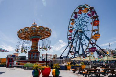 Kaliforniya 'daki Santa Cruz Sahili Boardwalk gündüz görüşü salıncaklı bir atlıkarınca, turuncu, sarı, mor gondollu bir dönme dolap, şemsiyeler, masalar ve okyanus kenarındaki ekicileri gösteriyor..