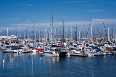 Monterey, Kaliforniya 'da Monterey Limanı sakin yansıtıcı su, gemi direkleri, rıhtım ormanları ve berrak gün ışığı rengi ve berraklığı üzerinde dolu yelkenli ve motor yatları gösteriyor..